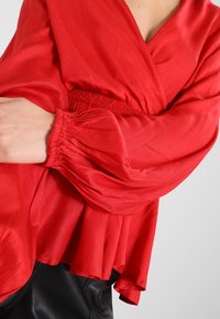 Rød bluse med indpakningsdesign, der har lange, samlede ærmer og en flared kant. Lavet af letvægtsstof med en glat tekstur.