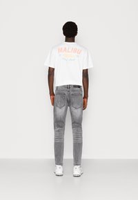 Vitt kortärmat t-shirt med "MALIBU BEACH 1990 Surf Shop" tryckt i pastellfärger, kombinerat med figurnära grå jeans och vita sneakers.