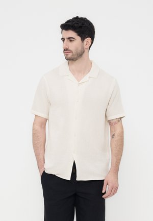 Homme aux cheveux foncés et à la barbe, portant une chemise crème à manches courtes boutonnée et un pantalon noir, regardant vers sa gauche, les mains dans les poches.