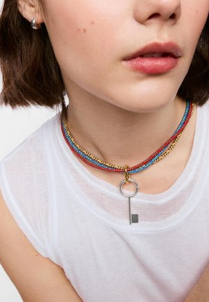 Zbliżenie osoby noszącej wielokolorowy naszyjnik choker z koralików z srebrnym wisiorkiem w kształcie klucza i srebrną kolczykiem-kołem.