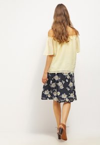 Haut jaune clair à épaules nues avec des bords volantés, associé à une jupe marine avec des motifs de marguerites blanches, et des chaussures à talons carrés marron.