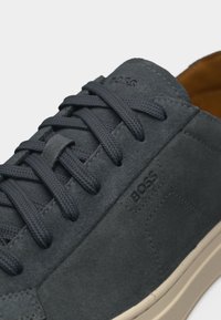 Σκούρο γκρι πάνινο αθλητικό παπούτσι με κορδόνια στο ίδιο χρώμα, ανάγλυφο λογότυπο "BOSS" στο πλάι και στη γλώσσα, ανοιχτό μπεζ σόλα, ταμπά εσωτερική επένδυση ορατή.