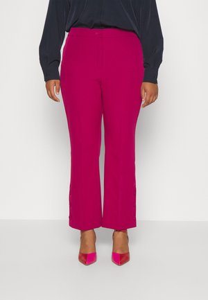 Vrouw draagt hooggetailleerde magentakleurige cropped broek met knoopdetails aan de zomen en bijpassende puntige hakken tegen een witte achtergrond.