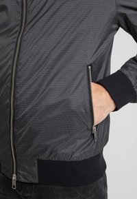 Svart bomberjacka med ett rutmönster, har en silverfärgad dragkedja och ribbade muddars. Materialet verkar slätt med en lätt glans.