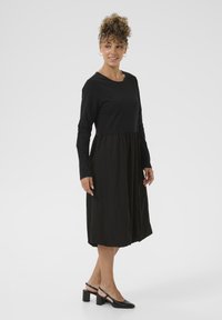 Crna haljina s dugim rukavima, uskim korzetom i A-oblika suknjom. Teksturirani materijal s glatkim gornjim dijelom. Uparena s crnim cipelama na visoku petu.