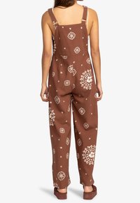 Bruine katoenen tuinbroek met een wit bloemmotief, verstelbare bandjes en een ontspannen pasvorm met wijde benen, ontworpen voor comfort en gemak.