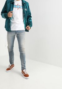 Teal satin bomberjacka, vit grafisk t-shirt med blå detaljer, ljus denimjeans och bruna mockasneakers med vita sulor.