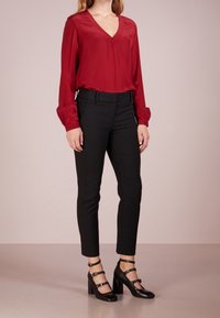 Žena v červené dlouhé blůze, černých upnutých kotníkových kalhotách a černých vysokých podpatcích se třemi pásky kolem kotníku.