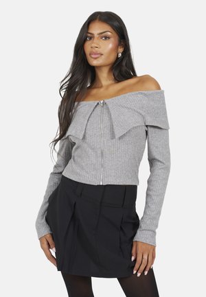 Donna con capelli lunghi e scuri che indossa un maglione grigio a coste con zip e scollatura a spalle scoperte e una minigonna nera plissettata contro sfondo bianco.