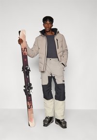 Snowboardista v béžové péřové bundě, šedém tričku a béžovo-černých lyžařských kalhotách, držící fialové lyže a s černými lyžařskými brýlemi.