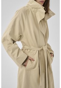 Moteris dėvi smėlio spalvos paltą su diržu, aukštu apykakliu ir rankomis kišenėse, stovinti priešais vientisą foną.