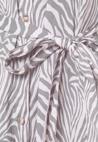 Tkanina se zebrovým vzorem v šedé a světle růžové barvě s mašlí v pase, má zapínání na knoflíky a hladkou texturu.