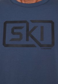 Tummansininen T-paita pehmeää kangasta, jossa on suuri musta "SKI" -logo sekä teksti "J. Lindeberg" oikeassa alakulmassa.