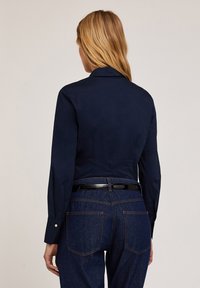 Camicia a maniche lunghe blu navy, in tessuto liscio, design con colletto, silhouette aderente, abbinata a jeans scuri di denim e a una cintura nera in pelle lucida.