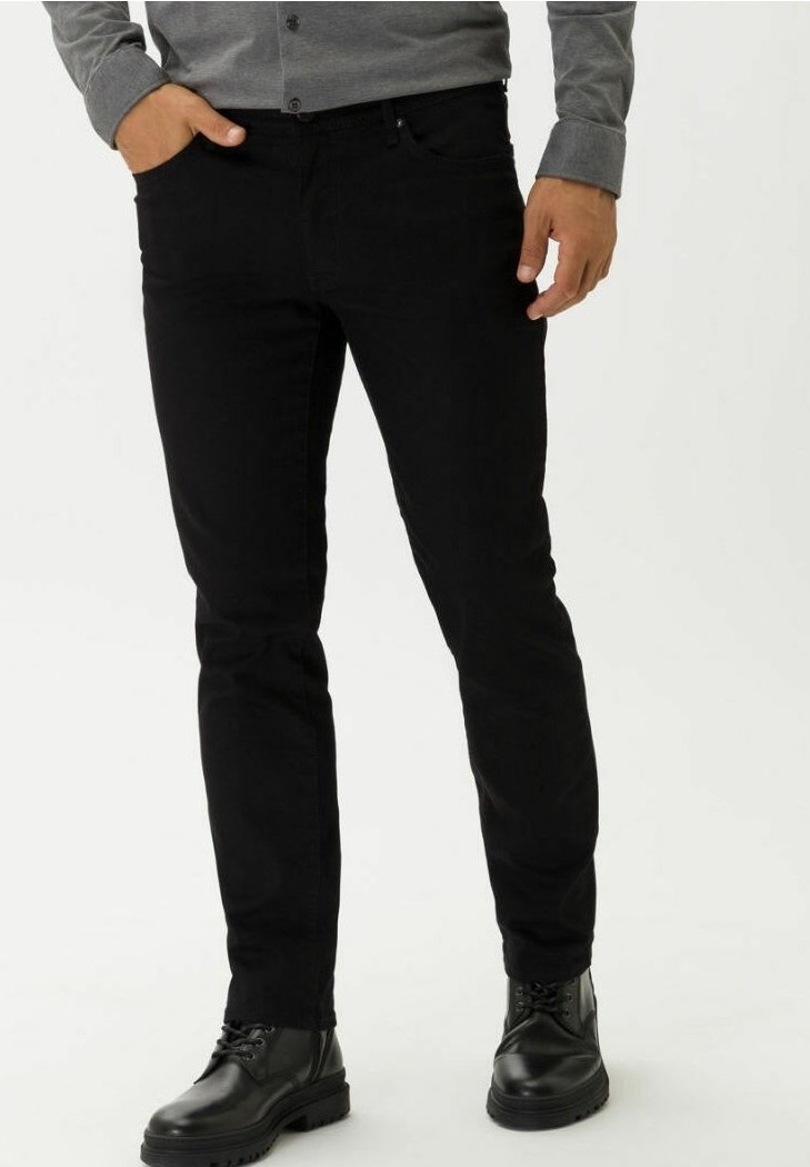 Homme portant un pantalon noir slim, des bottes noires à lacets, et une chemise grise à manches longues boutonnée, debout avec une main dans la poche.