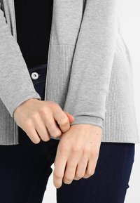 Světle šedý pletený cardigan s vroubkovanými rukávy, nošený přes černý top. Ruce drží manžety, což ukazuje texturu látky a detaily střihu.