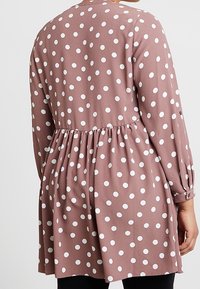 Hnědá šaty s puntíky, které mají dlouhé rukávy, nařasený pas a volný střih. Tkanina má hladkou strukturu s bílými kruhovými vzory.