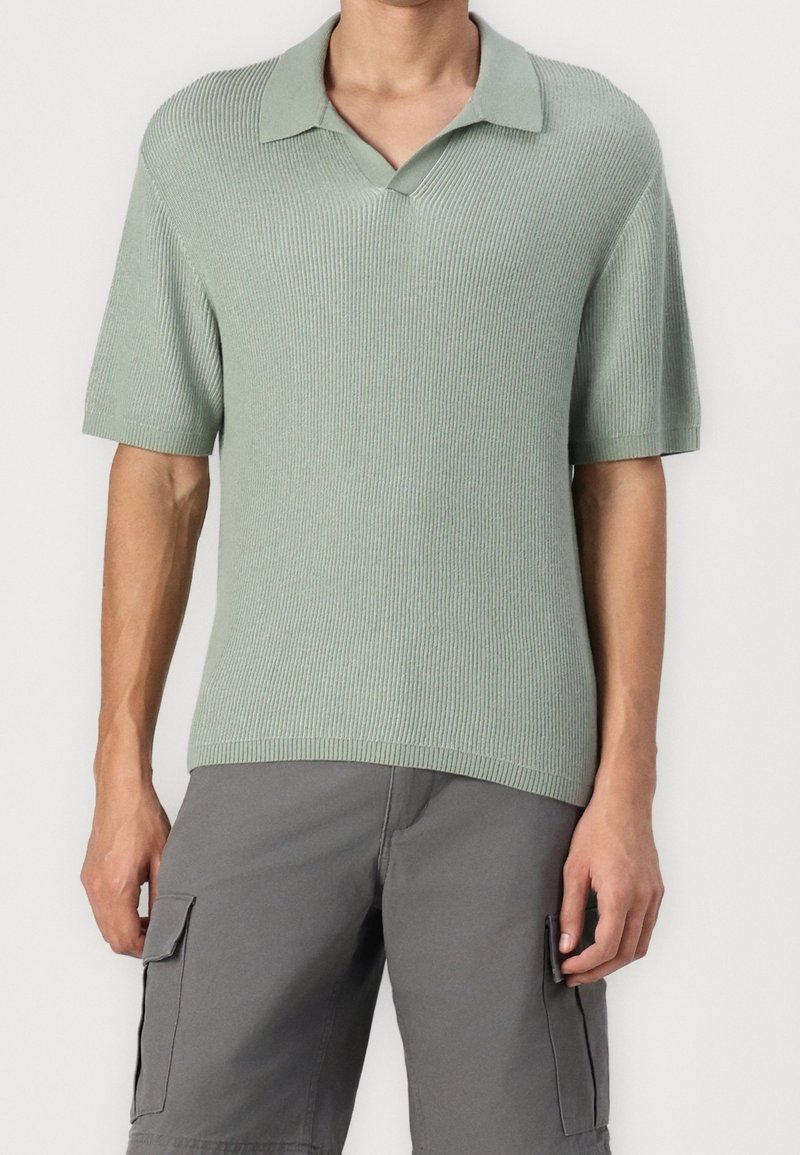 Man i en ljusgrön ribbad kortärmad pikétröja och grå cargoshorts med lockfickor, stående mot en neutral bakgrund.