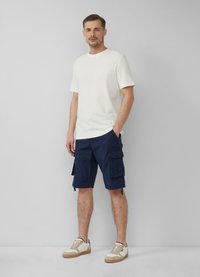 Witte t-shirt met korte mouwen, marineblauwe cargoshorts met zijzakken en beige sneakers met bruine zolen, staand tegen een lichtgrijze achtergrond.