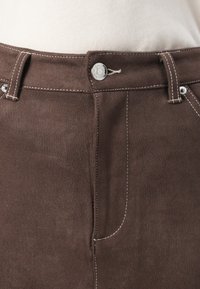 Brune suedebukser har en knappfrontlukkning, kontrasterende hvit søm, og en glatt tekstur med tilpasset passform og sidelommer.