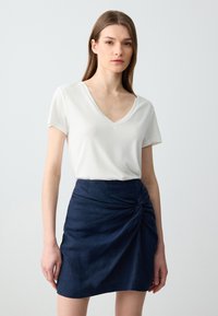 Weißes V-Ausschnitt T-Shirt mit kurzen Ärmeln, kombiniert mit einem dunkelblauen Jeansrock, der ein verdrehtes Knoten-Detail an der Taille aufweist.