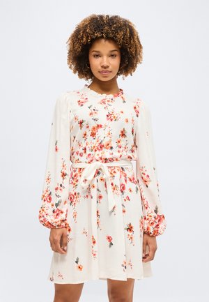 Νεαρή γυναίκα με σγουρά μαλλιά, φόρεμα μακρυμάνικο κρεμ με floral σχέδιο και δεμένο στη μέση, που στέκεται μπροστά σε απλό φωτεινό φόντο.