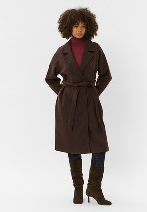 Femme aux cheveux bouclés portant un long manteau marron ceinturé sur un col roulé bordeaux, un pantalon noir et des bottines en daim marron à talons.