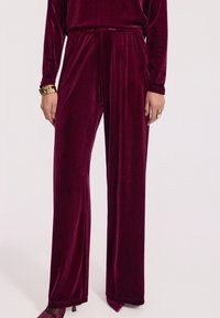 Burgundowe spodnie szerokie z weluru z elastycznym pasem, noszone z dopasowaną bluzką, czółenkami o spiczastym nosku, złotą bransoletką na nadgarstek i dużym pierścionkiem.
