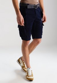 Muž v tmavě modrých cargo šortkách s bočními kapsami, šedým opaskem a béžovo-bílými teniskami Nike, stojící na světlém jednobarevném pozadí.