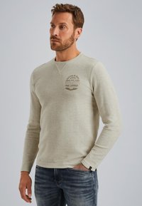 Light gray long-sleeve shirt with round neck, featuring a chest logo in dark brown and a textured fabric. Worn with blue jeans.