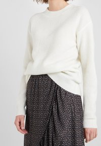 Witte gebreide trui met een ronde halslijn en lange mouwen, gecombineerd met een donkere gedrukte rok met een patroon van kleine, lichtgekleurde vormen.