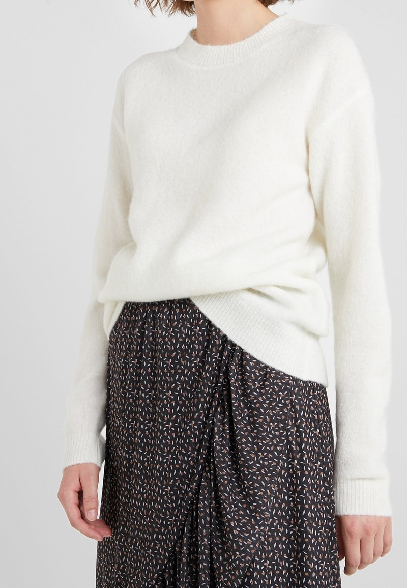 Witte gebreide trui met een ronde halslijn en lange mouwen, gecombineerd met een donkere gedrukte rok met een patroon van kleine, lichtgekleurde vormen.