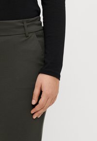Falda verde oliva con una textura suave, que presenta una cintura entallada y bolsillos laterales. Parte de una blusa negra de manga larga es visible.