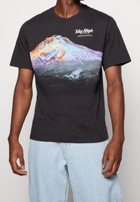 Svart t-shirt i bomull med en färgglad bergsgrafik i nyanser av lila, orange och blått, samt texten "Sky High" i vitt.