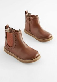 Bottes chelsea en cuir marron avec des panneaux élastiques sur les côtés, des bords festonnés, des perforations décoratives et une semelle cousue. Texture lisse et design décontracté.