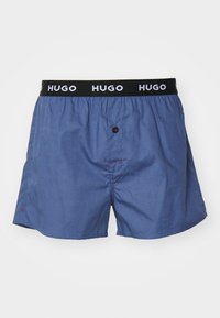 Boxeri din bumbac albastru cu un elastic negru, pe care este înscris logo-ul "HUGO". Include un nasture frontal și detalii de cusătură în contrast roșu.