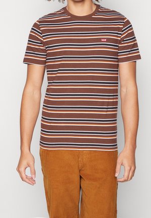 Personne portant un t-shirt Levi's marron à manches courtes rayé et un pantalon en velours côtelé marron clair, debout devant un fond uni.