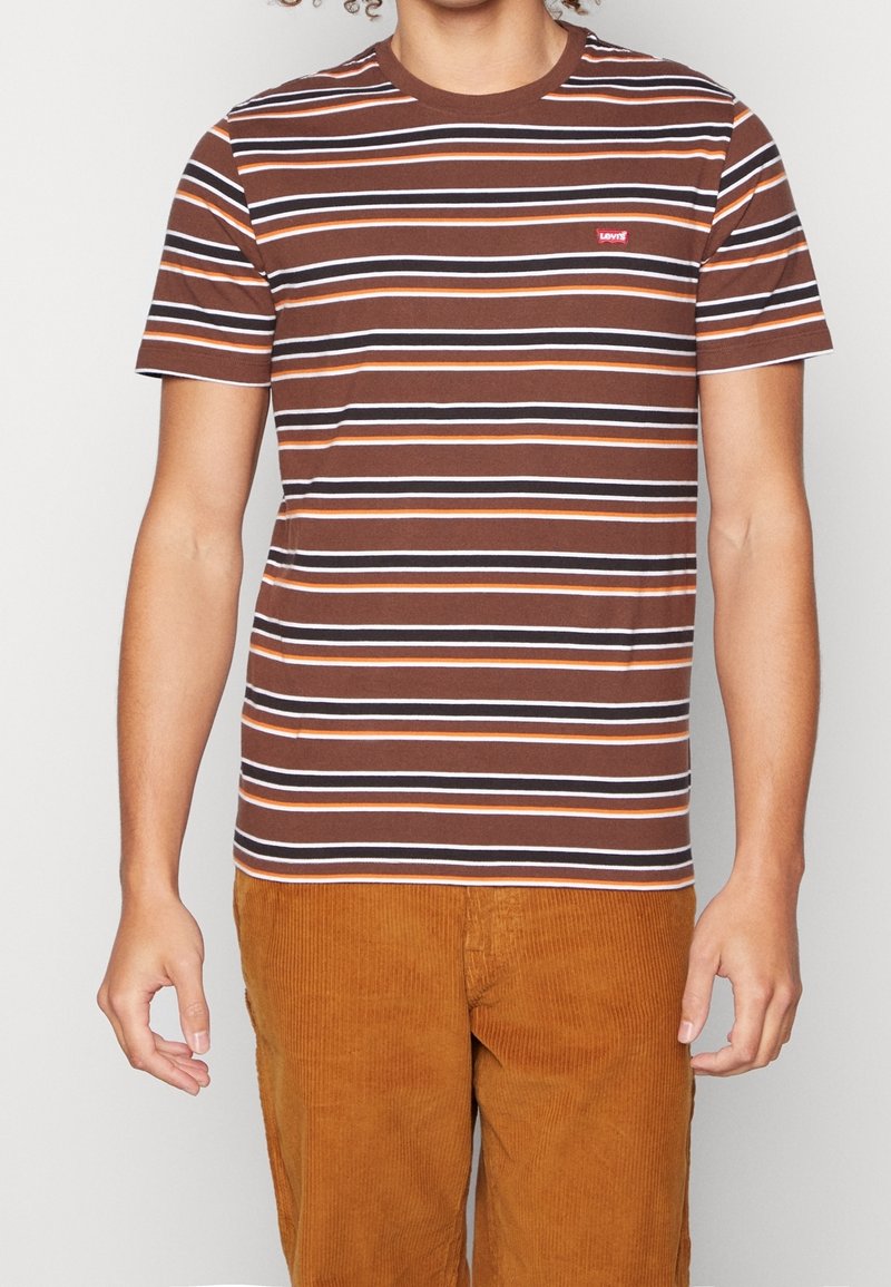 Personne portant un t-shirt Levi's marron à manches courtes rayé et un pantalon en velours côtelé marron clair, debout devant un fond uni.