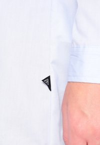 Světlomodrá košile s hladkou texturou, krátkými rukávy a černou trojúhelníkovou cedulkou s logem na bočním švu.