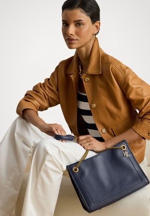 Γυναίκα με slicked-back μαλλιά φοράει καφέ δερμάτινο μπουφάν και άσπρο παντελόνι, κρατώντας ναυτικό τσάντα με χρυσή αλυσίδα και charm RL, καθισμένη σε κρέμα καναπέ.