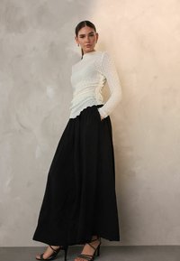 White, textured long-sleeve top with gathered detailing, paired with a flowing black maxi skirt. Model wears strappy black heeled sandals.