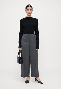 Vrouw die staat, gekleed in een zwarte lange mouwtop, grijze pinstripe wijde broek, zwarte hakken, en een zwarte handtas vasthoudend.