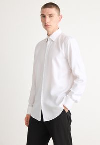 Chemise blanche à manches longues, boutonnée, avec un motif texturé, col classique et ourlet droit, associée à un pantalon noir.