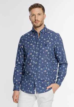 Mężczyzna ubrany w granatową koszulę zapinaną na guziki z wzorem plażowym i białe spodnie, stojący na tle jednolitego tła.