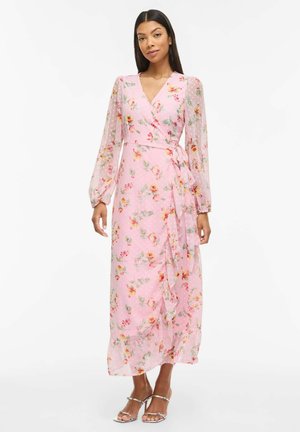 Kobieta ubrana w różową sukienkę kopertową z długim rękawem, ozdobioną kwiatowym wzorem i falbaną na dole, stoi przodem, mając na nogach srebrne sandały na obcasie.