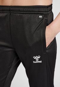 Melni sporta bikses, kas izgatavotas no gludas auduma, ar sānu kabatu, elastīgu jostu un baltu "hummel" logo tuvumā kabatai.