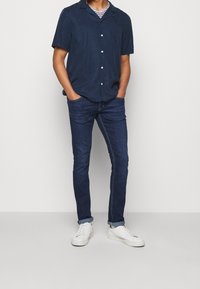 Camisa de manga corta azul marino con cuello y botones, combinada con jeans oscuros ajustados y zapatillas blancas. Diseño sencillo y limpio.