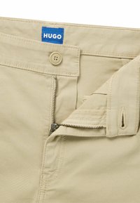 Pantaloni in cotone beige con zip e chiusura a bottone, dotati di etichetta blu "HUGO" in vita. Presentano una texture liscia e cuciture robuste.