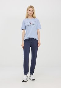 T-shirt azzurro chiaro con testo e design della bandiera, abbinato a pantaloni della tuta blu scuro. Scarpe da ginnastica bianche completano l'outfit. Stile minimalista.