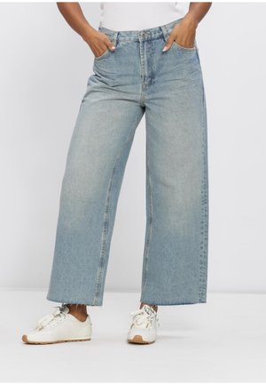 Mains dans les poches portant un jean large et court bleu clair et des baskets blanches à semelles en gomme sur fond blanc.